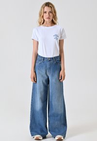 Witte t-shirt met blauwe bedrukte tekst, gecombineerd met lichtblauwe jeans met wijde pijpen. Soepele stof, ontspannen pasvorm en hoog ontwerp.