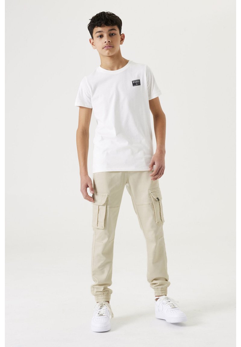 Chico adolescente de pie, vestido con camiseta blanca con pequeño logo en el pecho, pantalones cargo beige y zapatillas blancas, sobre fondo claro y liso.