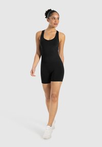 Monos de color negro ajustados con diseño sin mangas, escote redondeado y pantalones cortos. Hecho de un material suave y elástico. Lleva zapatillas blancas.