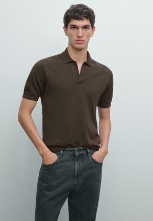 Jonge man met krullend haar, gekleed in een donkerbruine polo met korte mouwen en zwarte jeans, staand met handen in de zakken tegen een effen achtergrond.