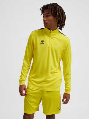 Jonge man draagt een felgeel sporttopje met lange mouwen en bijpassende korte broek met zwarte hummel-logo's, kijkt naar rechts.