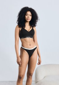 Crni grudnjak s V-izrezom, s brendiranim elastičnim pojasom. Usklađeni crni tangice s bijelim logom na pojasu.