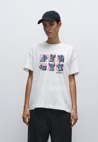 Hvid bomulds T-shirt med et farverigt grafisk motiv af en tegneseriefigur i seks positioner på en lyserød baggrund. Korte ærmer og afslappet pasform.