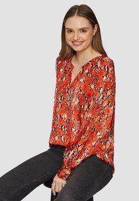 Rood en zwart slangenprint blouse met een V-hals en lange mouwen, gemaakt van gladde stof. Gecombineerd met donkere jeans zit het model op een kruk.