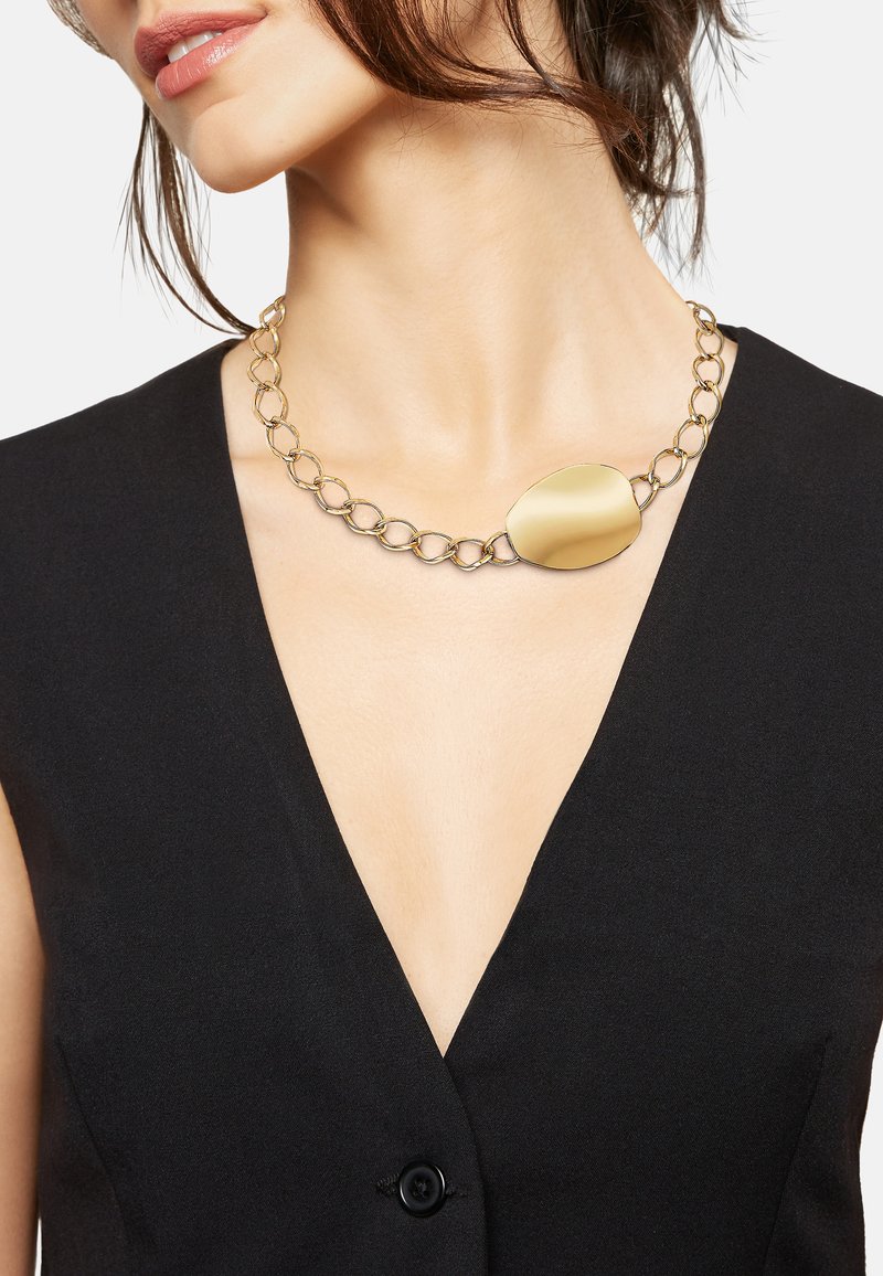Mujer que lleva un collar de cadena dorada con un gran colgante irregular y liso, combinado con una blusa sin mangas de cuello en V negra.