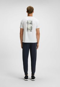 Hvid T-shirt med et grafisk print på ryggen, der viser abstrakte linjer og former; parret med navy atletisk bukser og sorte sneakers.
