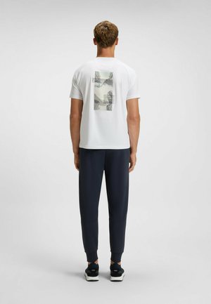 Weißes T-Shirt mit grafischem Druck auf der Rückseite, das abstrakte Linien und Formen zeigt; kombiniert mit navyblauen Sporthosen und schwarzen Turnschuhen.