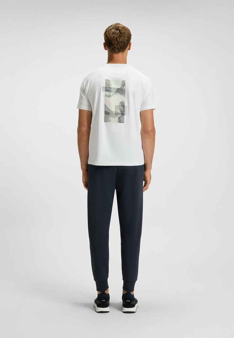 Hvid T-shirt med et grafisk print på ryggen, der viser abstrakte linjer og former; parret med navy atletisk bukser og sorte sneakers.