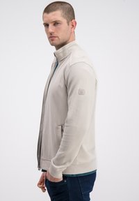 Jasno beżowa kurtka zapinana na zamek błyskawiczny z teksturowanymi rękawami, dwoma bocznymi kieszeniami i stójką. Wykonana z miękkiego, oddychającego materiału.