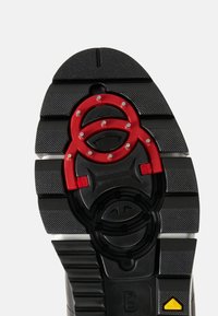 Black rubber sole with red accents and textured patterns. Features a circular design element in the center and a raised logo detail.