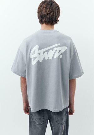 Osoba v voľnom sivom tričku s veľkým bielym štýlizovaným textom na chrbte, v kombinácii s vyblednutými tmavými džínami, stojaca na jednoduchom svetlom pozadí.