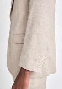 Beige Leinenanzugjacke mit tailliertem Fit, sichtbarer Textur und vier cremefarbenen Knöpfen am Ärmel. Keine sichtbaren Muster oder Akzente.