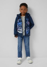 Junger Junge steht an der Wand und trägt einen marineblauen Kapuzenpullover mit blauem Tigerprint, ein gestreiftes Hemd, blaue Jeans und weiße Sneaker.