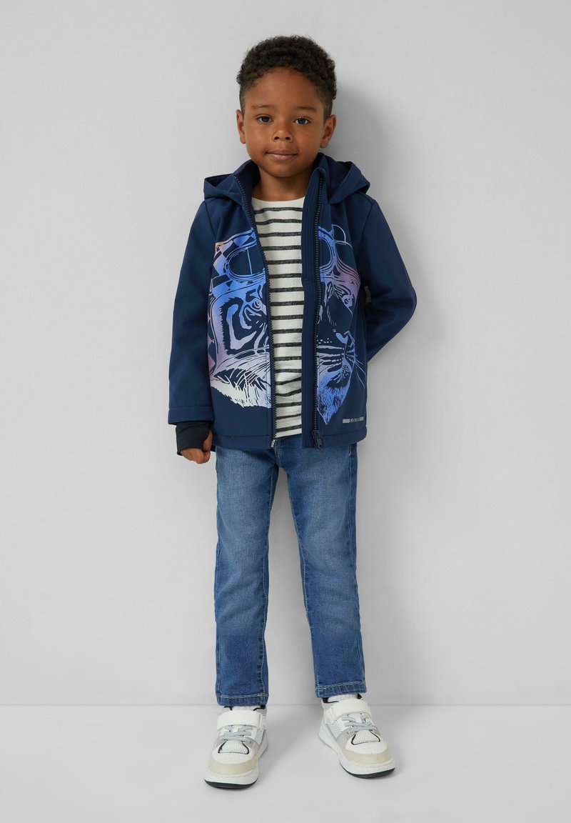 Junger Junge steht an der Wand und trägt einen marineblauen Kapuzenpullover mit blauem Tigerprint, ein gestreiftes Hemd, blaue Jeans und weiße Sneaker.