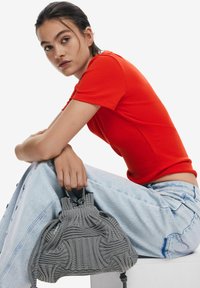 Graue gestreifte Tasche mit gerafftem Design und Kordelzugverschluss, kombiniert mit einem taillierten roten Oberteil und hellblauer Jeans.