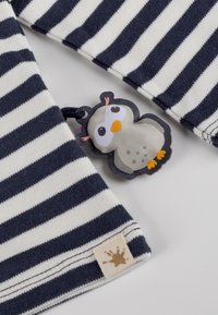 Gestreifter Stoff in Marineblau und Creme, mit einem Pinguin-förmigen Etikett. Das Etikett hat gestickte Augen, einen Schnabel und Polka-Dots. Enthält ein kleines Logopatch.