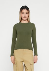 Top verde cu mâneci lungi, ajustat, cu decolteu rotund, confecționat dintr-un material moale și neted. Asortat cu pantaloni cargo bej deschis.