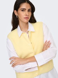 Geel fleecevest met een gestructureerd oppervlak, knoopsluiting en zakken, gedragen over een wit overhemd met kraag. Model poseert met gekruiste armen.