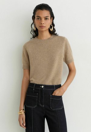 Vrouw met donker krullend haar draagt een beige korte mouwen trui, zwarte hoge taille broek met grote voorzakken, gouden creolen en een armband.