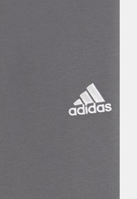 Sivi tekstil s glatkom površinom, s bijelim izvezenim Adidas logom u donjem kutu. Jednostavan, moderan dizajn.