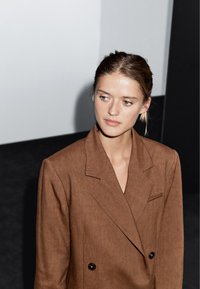 Brązowa lniana marynarka dwurzędowa z klapami i jedną kieszenią piersiową. Materiał ma teksturowany wygląd.