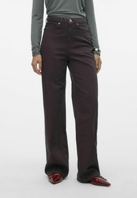 Jeans burgundy de pierna ancha con cintura alta, tela texturada y acentos de botones metálicos, combinados con una blusa gris de manga larga y zapatos rojos.