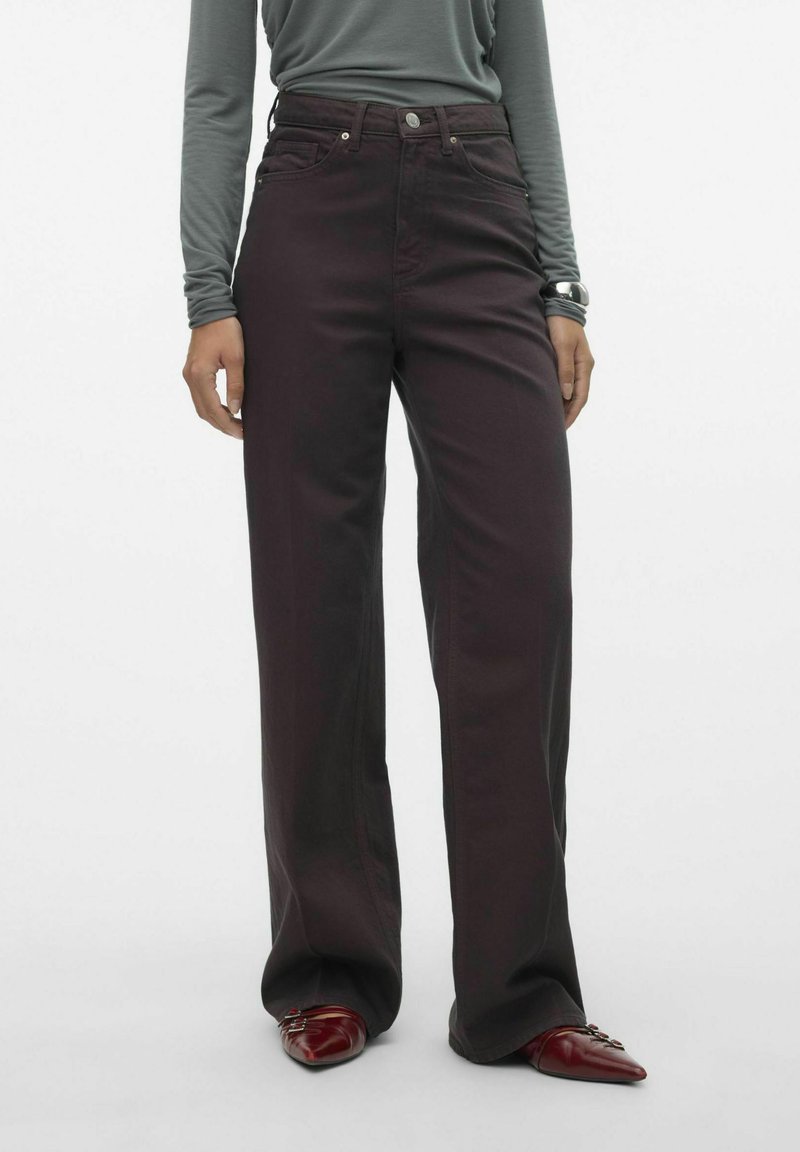 Jeans burgundy de pierna ancha con cintura alta, tela texturada y acentos de botones metálicos, combinados con una blusa gris de manga larga y zapatos rojos.