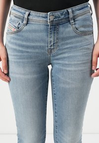 Light blue denim jeans feature a fitted design, five pockets, a metal button closure, and orange stitching accents. Smooth texture throughout.