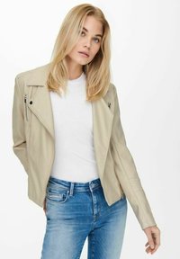 Beige skinnjakke i lær med asymmetrisk glidelås, skreddersydd passform og myk tekstur, brukt over en hvit t-skjorte og kombinert med blå jeans.