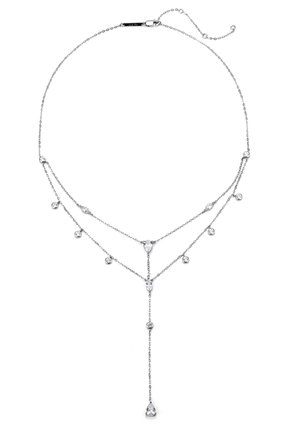 Kobieta nosząca warstwowy srebrny naszyjnik z małymi kamieniami, ubrana w biały top z wiązaniem na szyi, trzymająca łańcuszek naszyjnika blisko klatki piersiowej.