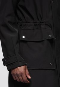 Czarna odzież wierzchnia o teksturowanym, gładkim wykończeniu. Posiada elastyczny pas, dużą kieszeń z patką oraz zatrzaski na mankietach.