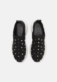 Fekete slip-on cipők fehér talppal, textúrázott felsőrésszel, amelyet fémes szegecsek mintázata díszít. Látható a DKNY logó.