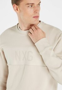 Beige sweatshirt met een textuurpaneel met geborduurde "NXG." Zacht materiaal, ribbelkraag en -manchetten, subtiele ontwerpelementen en een logo-accent.