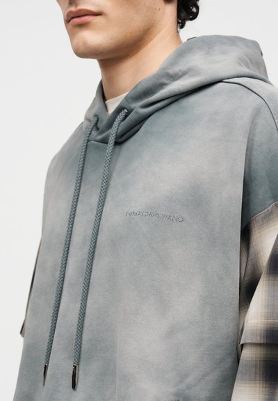 Κοντινό ενός ατόμου που φοράει γκρι φούτερ με κουκούλα και κορδόνια και καρό μανίκια, με μικρό κεντημένο κείμενο στο στήθος.