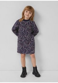 Junges Kind mit hellbraunen Haaren, das ein langärmliges lila Blumenkleid und schwarze geschnürte Stiefel trägt, steht vor einer schlichten Wand.