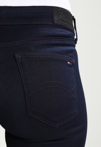 Jeans de mezclilla azul oscuro con un corte clásico, que presentan un bolsillo trasero con costuras sutiles y un pequeño detalle de logo en rojo, blanco y azul.
