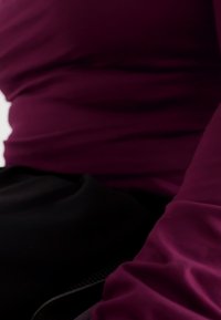 Μπορντό εφαρμοστό τοπ με μαλακή υφή, τοποθετημένο πάνω σε μαύρο ύφασμα. Το τοπ διαθέτει διακριτική πτώση και προσαρμοσμένη εφαρμογή γύρω από τη μέση.