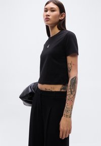 Μαύρο κοντομάνικο T-shirt από βαμβάκι, με μικρό λογότυπο στο στήθος. Το μοντέλο φοράει φαρδιά μαύρη παντελόνια και κρατά μία μαύρη τσάντα.