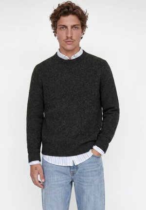 Homme aux cheveux bruns ondulés porte un pull vert foncé par-dessus une chemise blanche à rayures et un jean bleu clair, debout avec une main dans la poche.