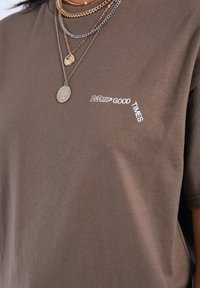 Camiseta de algodón marrón con el texto "Hazlo. Bien. Momentos." en blanco, con un corte relajado y mangas cortas, combinada con cadenas doradas.