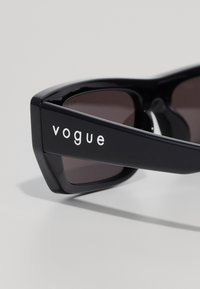 Juodos stačiakampės saulės akinių su tamsiais lęšiais, ant šventyklių rašoma "vogue" balta spalva, priešais šviesų foną.