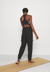 Musta urheilurintaliivit, joissa on selkäosassa leikkaus ja valkoinen raidallinen logo, yhdistettynä löysiin mustiin cargo-housuihin, joissa nilkkakuminauhat.