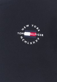 T-shirt di cotone blu navy con un logo ricamato circolare che presenta le scritte "NEW YORK", "TOMMY HILFIGER" e "MCMLXXXV", con dettagli in rosso, bianco e blu.