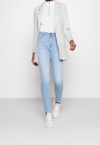 Svetle šedý dlouhý kardigan přes bílou mikinu na zip, doplněný o světlé modré skinny džíny a bílé tenisky. Hladké textury, moderní střih.