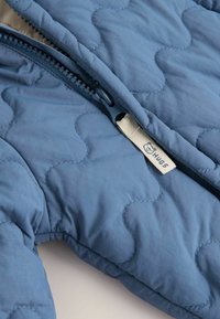 Gestepptes blaues Jacket mit einem marineblauen Reißverschluss und einem taupefarbenen Stoffetikett mit "HUGS"-Markierung. Weiche Textur mit Wellenmuster-Design.
