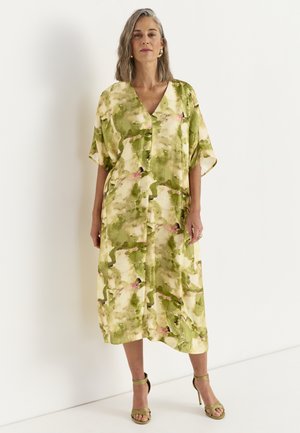 Kvinde iført løs, knælang kjole med grønt og beige abstrakt mønster, korte ærmer og V-hals, kombineret med grønne sandaler med stropper.