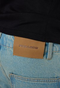 Črna majica z robom in modri jeans s rjavim usnjenim etiketom, šivano na hrbtni strani, z odtisnjenim napisom "PEGADOR."
