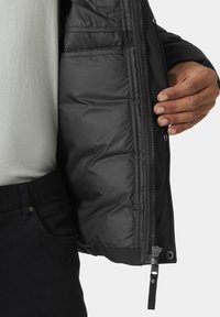 Chaqueta aislante negra con cierre de cremallera, bolsillos interiores y textura suave; parcialmente desabrochada, revelando una camiseta gris clara debajo.