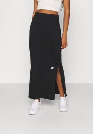 Donna con gonna lunga nera Nike con spacco laterale e sneakers bianche, in piedi davanti a uno sfondo chiaro e semplice.