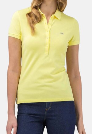 Donna che indossa una polo gialla a maniche corte con bottoni e jeans blu scuro, mostrata dal collo ai fianchi.